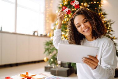 Noel ağacının arka planında elinde hediyeler, elinde tablet olan genç bir kadının arkadaşlarıyla görüntülü konuşması ya da ailesiyle görüntülü konuşması vardır. Noel çevrimiçi tatil. Evde tatil yap..
