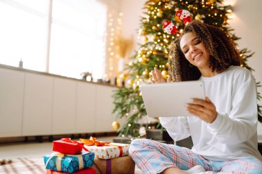Noel ağacının arka planında elinde hediyeler, elinde tablet olan genç bir kadının arkadaşlarıyla görüntülü konuşması ya da ailesiyle görüntülü konuşması vardır. Noel çevrimiçi tatil. Evde tatil yap..