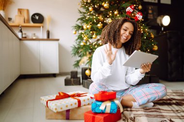 Noel ağacının arka planında elinde hediyeler, elinde tablet olan genç bir kadının arkadaşlarıyla görüntülü konuşması ya da ailesiyle görüntülü konuşması vardır. Noel çevrimiçi tatil. Evde tatil yap..