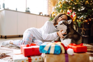 Güzel bir kadın yılbaşı ağacının yanında oturmuş köpeğiyle oynuyor ve eğleniyor. Genç bayan kırmızı Noel şapkasıyla köpeğine sarılıyor. Kış tatili Etrafta ışık.