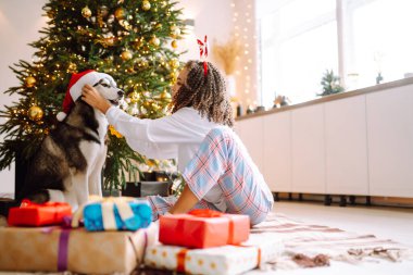 Güzel bir kadın yılbaşı ağacının yanında oturmuş köpeğiyle oynuyor ve eğleniyor. Genç bayan kırmızı Noel şapkasıyla köpeğine sarılıyor. Kış tatili Etrafta ışık.