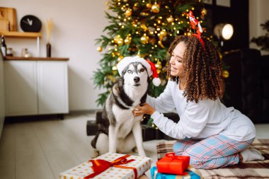 Güzel bir kadın yılbaşı ağacının yanında oturmuş köpeğiyle oynuyor ve eğleniyor. Genç bayan kırmızı Noel şapkasıyla köpeğine sarılıyor. Kış tatili Etrafta ışık.