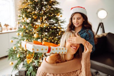 Şirin küçük bir kız, yakışıklı annesine bir hediye kutusu veriyor. Neşeli anne ve tatlı kızı birbirlerine hediye veriyorlar. Ebeveyn ve küçük çocuk içeride ağacın yanında eğleniyor. Sevgi dolu aile.