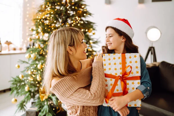Şirin küçük bir kız, yakışıklı annesine bir hediye kutusu veriyor. Neşeli anne ve tatlı kızı birbirlerine hediye veriyorlar. Ebeveyn ve küçük çocuk içeride ağacın yanında eğleniyor. Sevgi dolu aile.