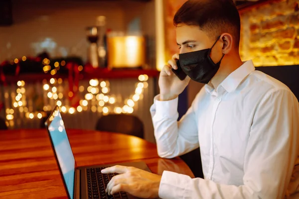 医療用フェイスマスクをした若者がノートパソコンで働き 冬休みには自宅の電話で話す 流行コロナウイルスの拡散を防ぐという概念 リモートワーク — ストック写真