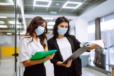 Koruyucu yüz maskesi takmış iki iş arkadaşı ofis koridorunda işle ilgili konuları tartışıyor. Karantina şehrindeki salgın sırasında ofiste çalışan iki kadın ofis çalışanı.. 