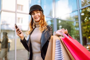 Telefonunu kullanarak alışveriş torbalarıyla gülümseyen kadın. Şehir caddesinde alışveriş yapan genç bir kadın. Satın almalar, kara cuma, indirimler, satış konsepti. Çevrimiçi alışveriş kavramı.