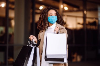Mağazanın yakınında alışveriş yaptıktan sonra koruyucu maske takan güzel Afro-Amerikan kadın portresi. Coronavirus Covid-19 salgını sırasında alışveriş. Satışlar ve İndirim, Perakende, Satın Alım.