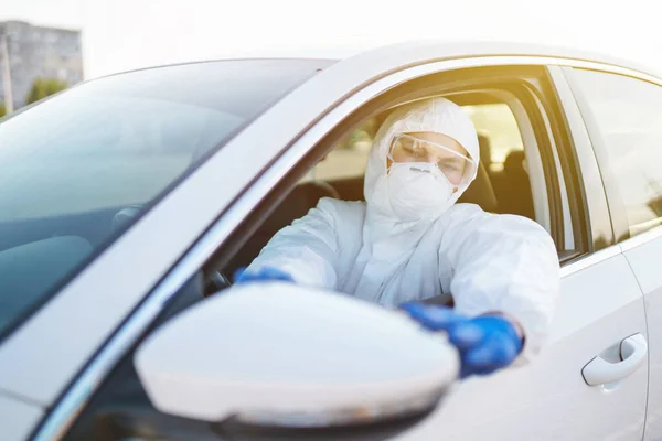 Man in virus protective suit and mask driving car.  Man wearing protective hazmat rides in the car. The concept of preventing the spread of the epidemic, pandemic in quarantine city. Covid -19.