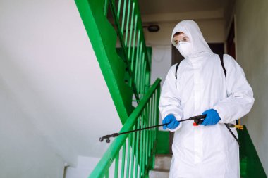 Koruyucu giysi uzmanı bir konut binasının girişini dezenfekte ediyor. Koruyucu koruyucu koruyucu giysili ve maskeli bir adam salgının yayılmasını engellemek için dezenfektan püskürtüyor. Covid 2019. 