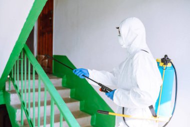 Koruyucu giysi uzmanı bir konut binasının girişini dezenfekte ediyor. Koruyucu koruyucu koruyucu giysili ve maskeli bir adam salgının yayılmasını engellemek için dezenfektan püskürtüyor. Covid 2019. 
