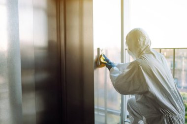 Asansörü dezenfekte etmek. Koruyucu kıyafetli ve maskeli bir adam asansörün dezenfektan düğmelerini spreyliyor. COVID-19 hastalığını koruma amaçlı. Temizlik kavramı. 