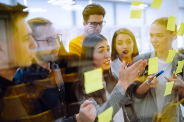 Genç iş adamları şirket projesinde birlikte beyin fırtınası yapıyorlar. Yapışkan notlarla strateji görevleri yazıyorlar. Cam duvarın arkasında duruyorlar..