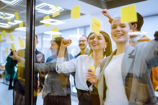 Young business team people brainstorm on corporate project together write strategy tasks on post in sticky notes stand behind glass wall, diverse staff group planning management concept.