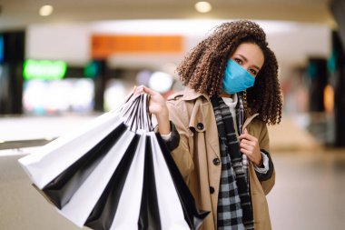 Koruyucu steril tıbbi maskeli Afro Amerikalı kadın alışveriş merkezinden alışveriş yapıyor, alışverişe çıkıyor. Bahar alışverişi. Satın alma, alışveriş, yaşam tarzı konsepti. 