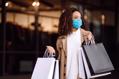 Alışveriş merkezinin yakınında alışveriş yaptıktan sonra yüzünde koruyucu steril tıbbi maske takan genç bir kadın. Bahar alışverişi. Satın alma, alışveriş, yaşam tarzı konsepti. 