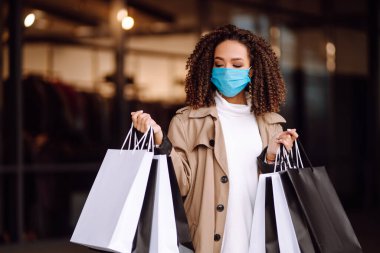 Alışveriş merkezinin yakınında alışveriş yaptıktan sonra yüzünde koruyucu steril tıbbi maske takan genç bir kadın. Bahar alışverişi. Satın alma, alışveriş, yaşam tarzı konsepti. 