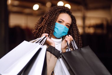 Alışveriş merkezinin yakınında alışveriş yaptıktan sonra yüzünde koruyucu steril tıbbi maske takan genç bir kadın. Bahar alışverişi. Satın alma, alışveriş, yaşam tarzı konsepti. 