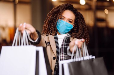 Alışveriş merkezinin yakınında alışveriş yaptıktan sonra yüzünde koruyucu steril tıbbi maske takan genç bir kadın. Bahar alışverişi. Satın alma, alışveriş, yaşam tarzı konsepti. 