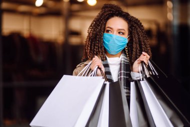 Alışveriş merkezinin yakınında alışveriş yaptıktan sonra yüzünde koruyucu steril tıbbi maske takan genç bir kadın. Bahar alışverişi. Satın alma, alışveriş, yaşam tarzı konsepti. 