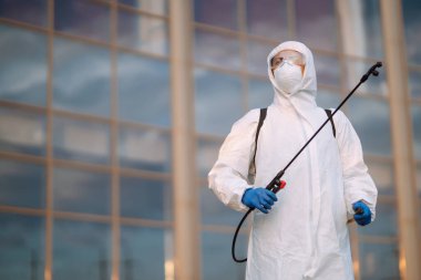 Tehlikeli madde giysisi giyen, koruyucu eldivenli ve gözlüklü bir adam dezenfekte cihazı kullanıyor. Covid-2019. Karantina şehrinde küresel salgın virüsünün yayılmasını önlemek.