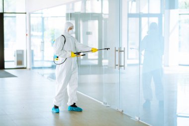 Tehlikeli madde giysisi giyen, koruyucu eldivenli ve gözlüklü bir adam ofiste dezenfekte cihazı kullanıyor. COVID-19 hastalığının korunması.