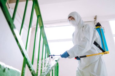 Koruyucu eldiven ve gözlük takan tehlikeli madde giysisi giyen bir adam bir apartmanın girişini dezenfekte ediyor. Covid 2019.