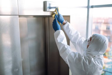 COVID-19 'u önlemek için asansörün temizlenmesi ve dezenfekte edilmesi. Tehlikeli madde giysili bir adam, koruyucu eldivenler ve asansörün dezenfekte düğmeleri. Covid 2019.