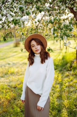 Çiçekli bahar parkında poz veren şapkalı, gülümseyen kadın. Rahatlama, seyahat, özgürlük ve bahar tatili kavramı. Moda tarzı.