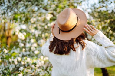 Çiçekli bahar parkında poz veren şapkalı, gülümseyen kadın. Rahatlama, seyahat, özgürlük ve bahar tatili kavramı. Moda tarzı.
