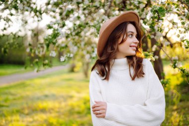 Çiçekli bahar parkında poz veren şapkalı, gülümseyen kadın. Rahatlama, seyahat, özgürlük ve bahar tatili kavramı. Moda tarzı.