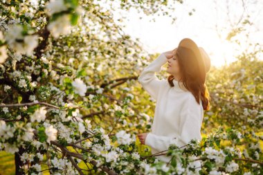 Çiçekli bahar parkında poz veren şapkalı, gülümseyen kadın. Rahatlama, seyahat, özgürlük ve bahar tatili kavramı. Moda tarzı.