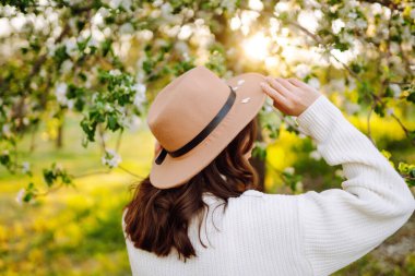 Çiçekli bahar parkında poz veren şapkalı, gülümseyen kadın. Rahatlama, seyahat, özgürlük ve bahar tatili kavramı. Moda tarzı.