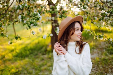 Çiçekli bahar parkında poz veren şapkalı, gülümseyen kadın. Rahatlama, seyahat, özgürlük ve bahar tatili kavramı. Moda tarzı.