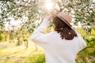 Çiçekli bahar parkında poz veren şapkalı, gülümseyen kadın. Rahatlama, seyahat, özgürlük ve bahar tatili kavramı. Moda tarzı.