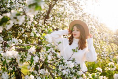 Çiçekli bahar parkında poz veren şapkalı, gülümseyen kadın. Rahatlama, seyahat, özgürlük ve bahar tatili kavramı. Moda tarzı.