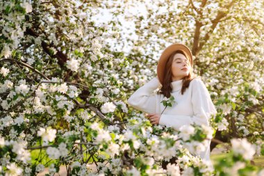 Çiçekli bahar parkında poz veren şapkalı, gülümseyen kadın. Rahatlama, seyahat, özgürlük ve bahar tatili kavramı. Moda tarzı.