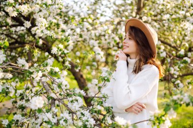 Çiçekli bahar parkında poz veren şapkalı, gülümseyen kadın. Rahatlama, seyahat, özgürlük ve bahar tatili kavramı. Moda tarzı.