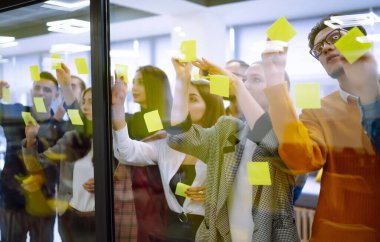 Bir grup modern insan proje üzerinde birlikte çalışıyor. İş arkadaşları cam duvara yapışkan bir not yazıyorlar ve iş arkadaşları ofiste bekliyor. Girişim işi. Planlama, analiz.