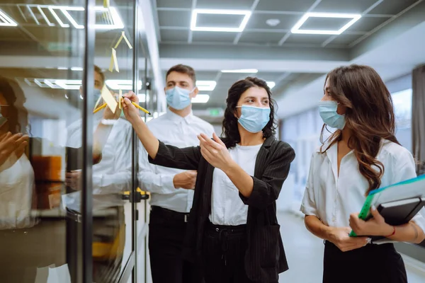 Business colleagues  in protective face mask writing on sticky note on glass wall with coworkers standing by in office. Startup Business. Planning, analysis. Covid-2019.