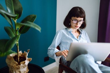 Genç kadın serbest yazar evdeki oturma odasında modern kanepeye oturup dizüstü bilgisayar yazıyor. Eğitim çevrimiçi. Karantina karantinasında evden çalışıyor. Sosyal uzaklık. Kişisel İzolasyon.