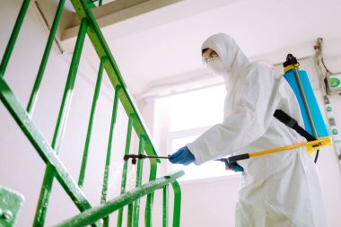 Korunaklı kıyafet giyen bir adam karantina şehrinde koronavirüsün yayılmasını önlemek için sprey kimyasallarla bir apartmanın girişini dezenfekte ediyor. Covid -19. Temizlik kavramı.