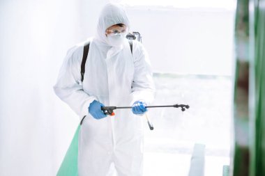Korunaklı kıyafet giyen bir adam karantina şehrinde koronavirüsün yayılmasını önlemek için sprey kimyasallarla bir apartmanın girişini dezenfekte ediyor. Covid -19. Temizlik kavramı.