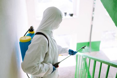 Korunaklı kıyafet giyen bir adam karantina şehrinde koronavirüsün yayılmasını önlemek için sprey kimyasallarla bir apartmanın girişini dezenfekte ediyor. Covid -19. Temizlik kavramı.