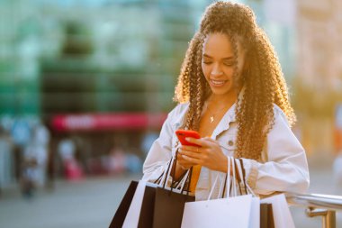 Alışveriş çantalı genç bir kadın sokakta cep telefonu kullanıyor. Bahar stili. Tüketim, satış, alım, alışveriş, yaşam tarzı konsepti.