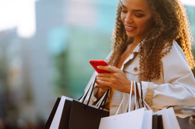 Alışveriş çantalı genç bir kadın sokakta cep telefonu kullanıyor. Bahar stili. Tüketim, satış, alım, alışveriş, yaşam tarzı konsepti.