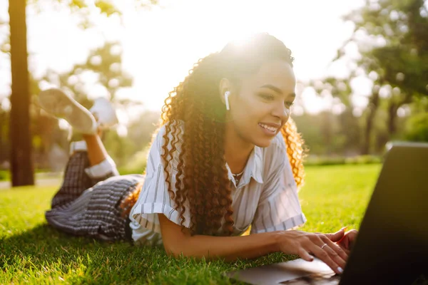 Young woman freelancer sitting on green grass with laptop. Education online. Happy woman with wireless headphones calling on laptop, talk by webcam, video conference.