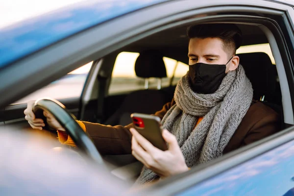 保護医療マスクの男性ドライバーは車の中で座って携帯電話を使用しました コロナウイルスの発生中に街の路上で運転者 Covid — ストック写真