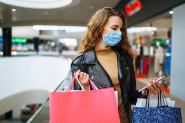 Modern mağaza merkezinde çok renkli çantaları olan koruyucu tıbbi maskeli şık bir kadın. Telefonunu kullanan genç bir kadın. Tüketim keyfi, hediye alışverişi, tatil. Çevrimiçi alışveriş kavramı.