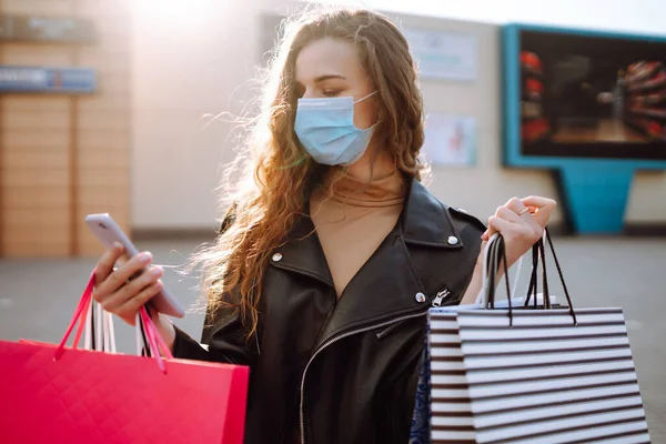 彼女の携帯電話を使用して買い物袋と保護医療用マスクの笑顔の女性 街の通りで買い物をした若い女性 オンラインショッピングのコンセプト コロナウイルス流行のショッピング — ストック写真